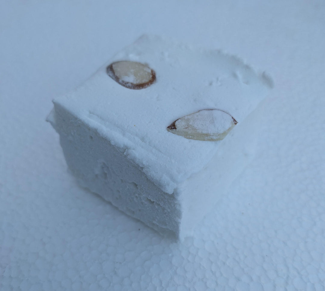 A single almond cake marshmallow with slivers of almonds on top, displayed on a clean, white background. The marshmallow is cut to approximately 2" x 2" x 1.5+", showcasing its golden brown, toasted exterior and the delicate almond slivers. The simplicity of the white background emphasizes the artisanal quality and delightful appearance of the marshmallow.