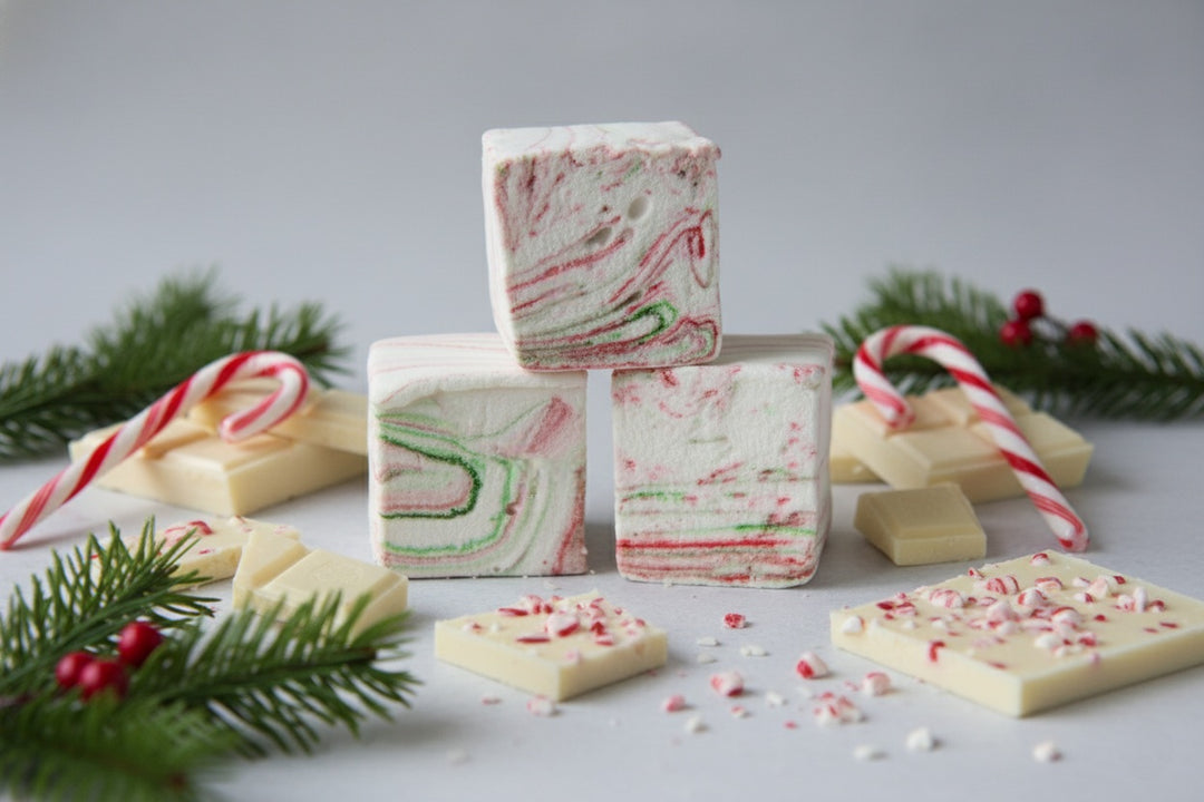 Stack of three White Chocolate Peppermint Bark marshmallows by The Toasty S’more, with crushed candy canes in the background, white chocolate pieces, and festive Christmas branches on a bright white backdrop. Handcrafted with beef gelatin and organic marshmallow root for a corn-syrup free, clean-label holiday treat.