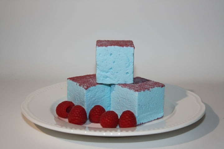 Three blue raspberry gourmet marshmallows made with beef gelatin and organic marshmallow root, stacked vertically to showcase their vibrant blue color and red sugar topping. Fresh red raspberries placed nearby enhance the clean-label, corn syrup-free presentation designed for bold flavor and festive gifting.