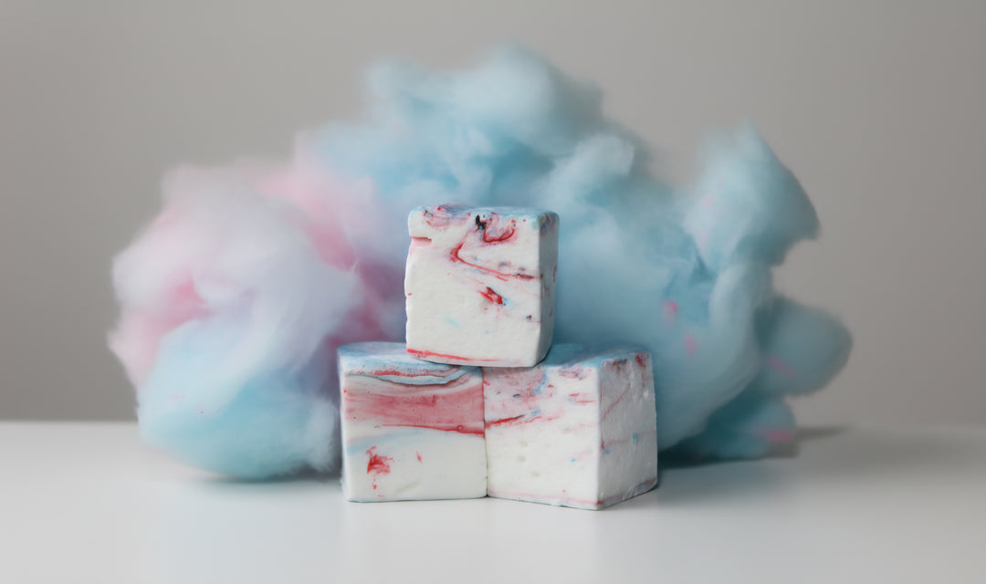 Three cotton candy gourmet marshmallows made with beef gelatin and organic marshmallow root, stacked vertically to highlight their pink and blue swirl. Fluffy cotton candy in the background enhances the whimsical, carnival-inspired styling. Clean-label, corn syrup-free marshmallows crafted for nostalgic flavor and vibrant gifting.