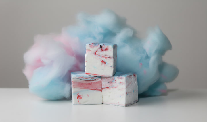 Three cotton candy gourmet marshmallows made with beef gelatin and organic marshmallow root, stacked vertically to highlight their pink and blue swirl. Fluffy cotton candy in the background enhances the whimsical, carnival-inspired styling. Clean-label, corn syrup-free marshmallows crafted for nostalgic flavor and vibrant gifting.