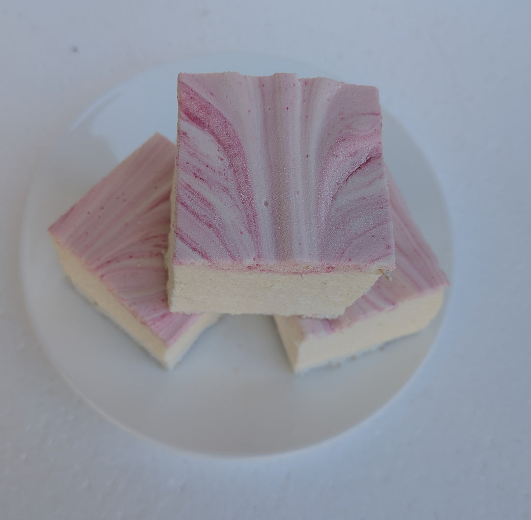 Top‑down view of three handcrafted strawberry cheesecake marshmallows made with natural colors, halal‑friendly beef gelatin, organic marshmallow root, and clean‑label ingredients, arranged on a white plate and showing their soft, fluffy texture with rich cheesecake flavor and strawberry accents.