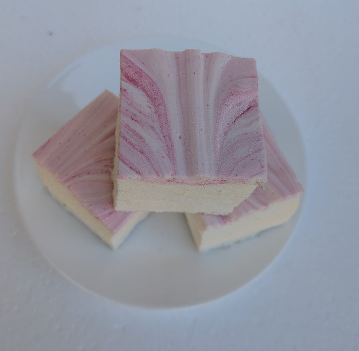 Top‑down view of three handcrafted strawberry cheesecake marshmallows made with natural colors, halal‑friendly beef gelatin, organic marshmallow root, and clean‑label ingredients, arranged on a white plate and showing their soft, fluffy texture with rich cheesecake flavor and strawberry accents.