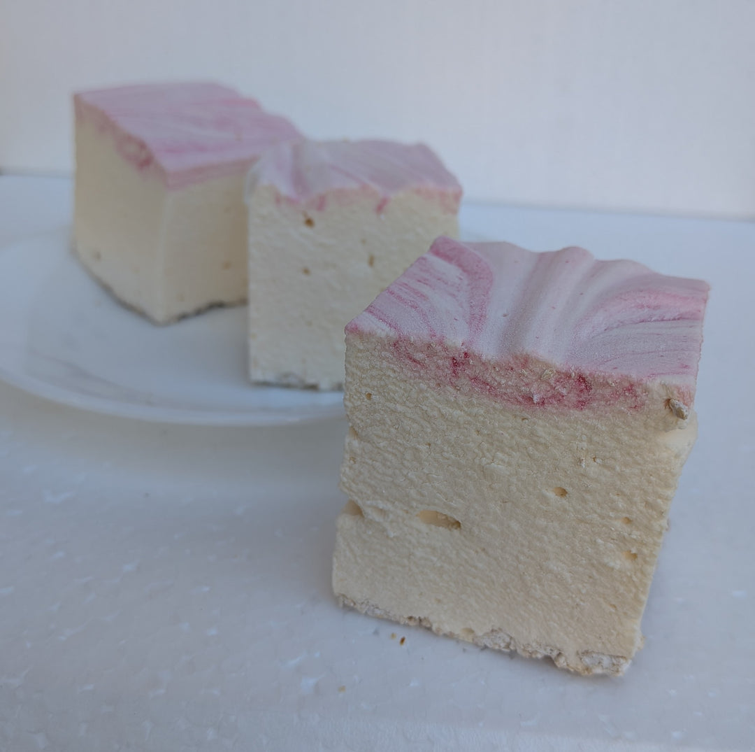 Three handcrafted strawberry cheesecake marshmallows made with natural colors, halal‑friendly beef gelatin, organic marshmallow root, and clean‑label ingredients, arranged with one marshmallow in front to highlight its soft, fluffy texture and rich cheesecake flavor with strawberry accents.