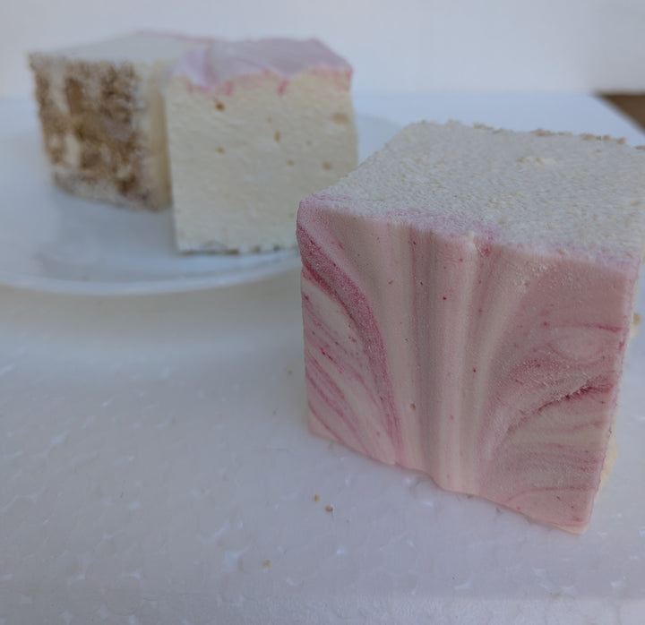 Three handcrafted strawberry cheesecake marshmallows made with natural colors, beef gelatin, organic marshmallow root, and clean‑label ingredients, arranged with one marshmallow in front and a graham cracker visible behind, highlighting their soft, fluffy texture and strawberry‑cheesecake flavor.