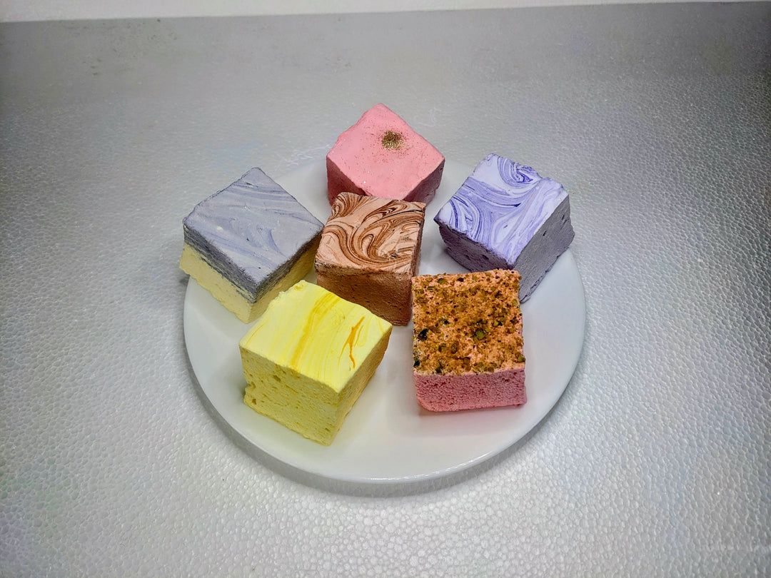 An elegant white plate displaying six varieties of gourmet marshmallows: lavender & honey, rose pistachio, creamy taro, champagne & sugar, lemon pound cake, and dulce de leche. Each marshmallow is handcrafted and generously sized
