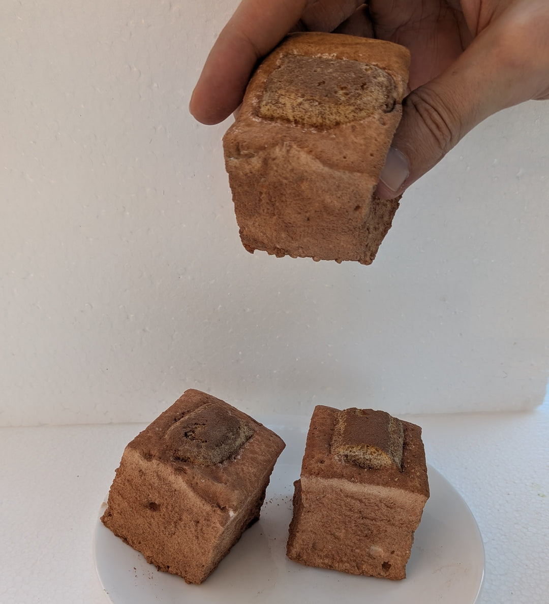 Hand holding a cube of Tiramisu Gourmet Marshmallows above three similar cubes on a white surface.
