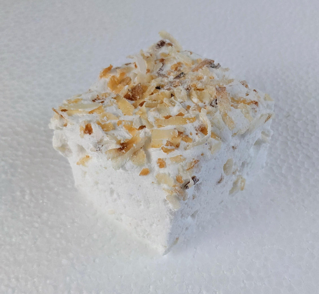 A single toasted coconut marshmallow made with beef gelatin, organic marshmallow root, and no corn syrup, displayed against a clean white background. Approximately 2" x 2" x 2", the marshmallow features a soft, fluffy texture and golden coconut flakes that highlight its handcrafted quality and clean-label ingredients.