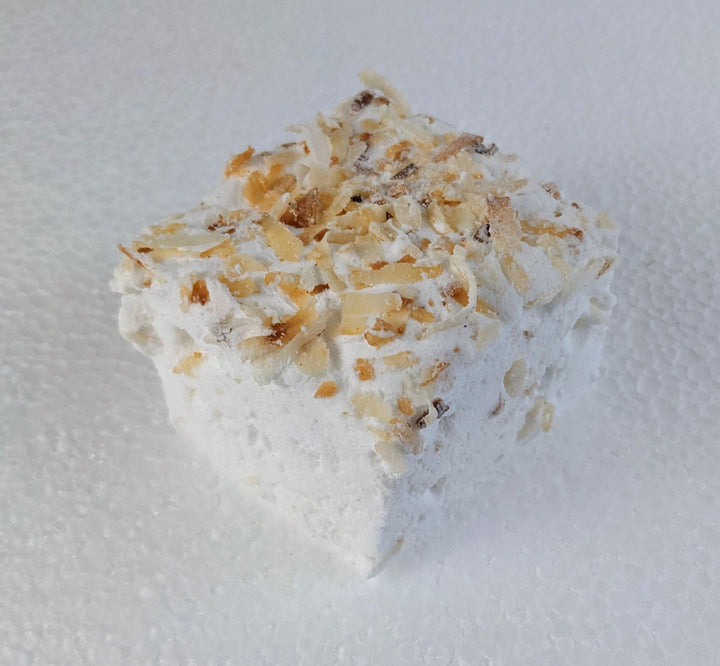 A single toasted coconut marshmallow made with beef gelatin, organic marshmallow root, and no corn syrup, displayed against a clean white background. Approximately 2" x 2" x 2", the marshmallow features a soft, fluffy texture and golden coconut flakes that highlight its handcrafted quality and clean-label ingredients.