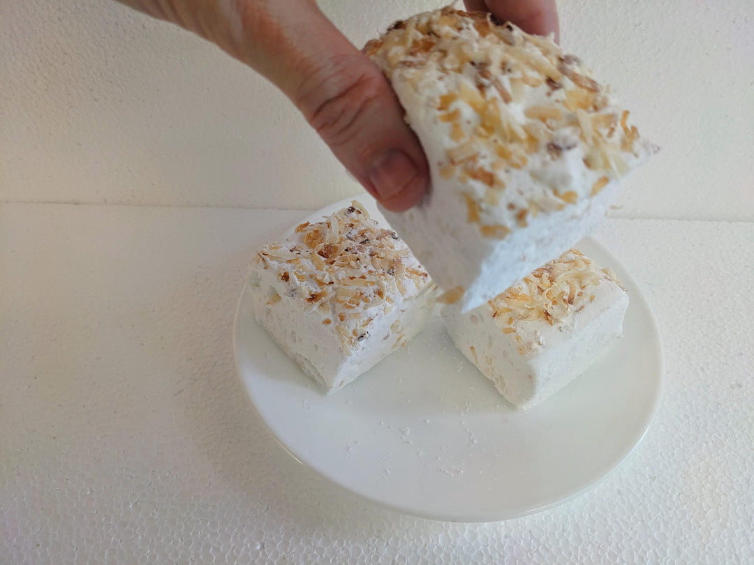 Handcrafted toasted coconut marshmallows made with beef gelatin, organic marshmallow root, and no corn syrup, displayed against a clean white background. Each marshmallow measures approximately 2" x 2" x 2", featuring a soft, fluffy texture and golden coconut flakes. A hand is shown picking up one marshmallow, emphasizing its pillowy interior and artisanal quality.