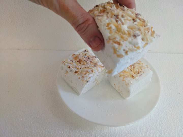 Handcrafted toasted coconut marshmallows made with beef gelatin, organic marshmallow root, and no corn syrup, displayed against a clean white background. Each marshmallow measures approximately 2" x 2" x 2", featuring a soft, fluffy texture and golden coconut flakes. A hand is shown picking up one marshmallow, emphasizing its pillowy interior and artisanal quality.