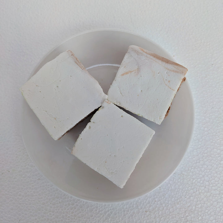 From a top-down view, three golden Butterbeer marshmallows rest on a clean, white plate, with one slightly in front, revealing its pillowy texture. Their extra-large 2-inch cubes showcase a butterscotch-vanilla essence, handcrafted in the USA with premium gelatin and no corn syrup for the perfect melt-in-your-mouth experience. Ideal for s’mores, desserts, or gourmet gifting, these marshmallows bring a magical touch to any occasion.
