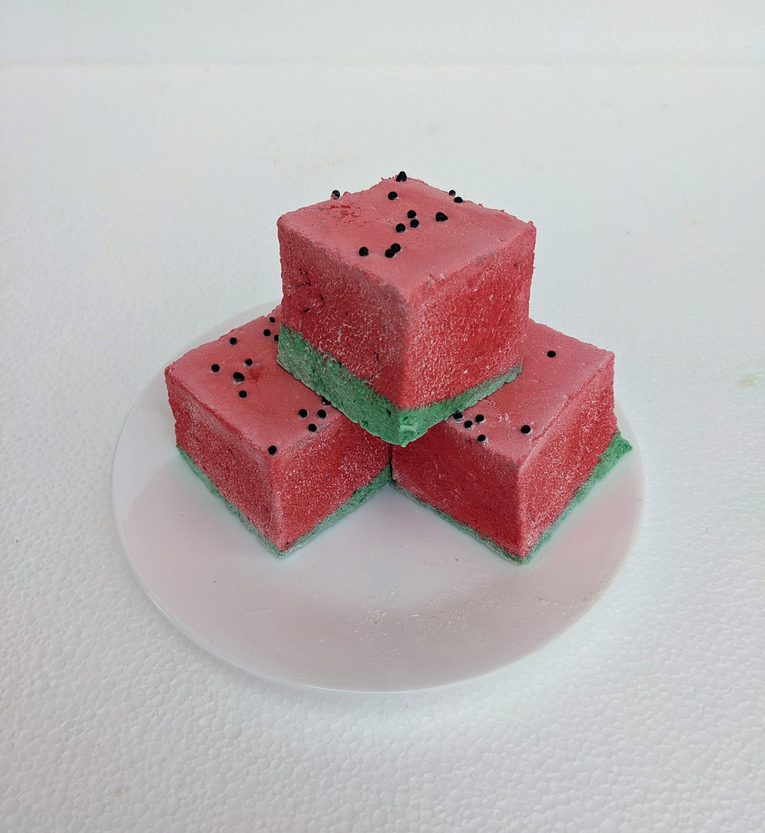 A close-up view of three watermelon-inspired gourmet marshmallows arranged on a white plate. Each marshmallow features a green base and a red top, designed to resemble a juicy watermelon slice. Tiny black sprinkles are scattered across the red layer, mimicking watermelon seeds. The bright colors contrast beautifully against the clean, white plate, emphasizing their fluffy texture and handcrafted quality.