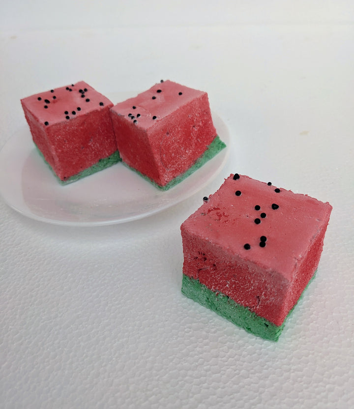 A close-up view of three of the best fruity marshmallows, inspired by watermelon, arranged on a white plate with one marshmallow in front and two in the back. Each marshmallow features a green base and a red top, designed to resemble a juicy watermelon slice. Tiny black sprinkles are scattered across the red layer, mimicking watermelon seeds. The bright colors contrast beautifully against the clean, white plate, emphasizing their fluffy texture and handcrafted quality. A fun and delicious treat for fruit-fl