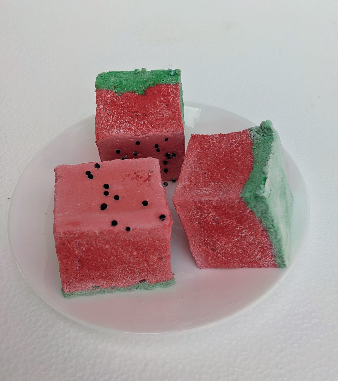 A close-up toppled arrangement of three watermelon-inspired gourmet marshmallows, handcrafted and made in the USA by The Toasty Smore, resting on a white plate. The marshmallows feature a green base and a red top, designed to resemble a juicy watermelon slice. Tiny black sprinkles are scattered across the red layer, mimicking watermelon seeds. Some marshmallows lean against each other, showcasing their fluffy texture and vibrant colors, making them one of the best fruity marshmallow treats for a fun and del