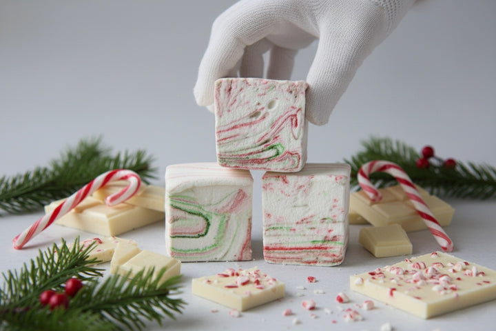 White Chocolate Peppermint Bark marshmallows by The Toasty S’more, held in a gloved hand with crushed candy canes, white chocolate pieces, and festive Christmas branches in the background. Handcrafted with beef gelatin and organic marshmallow root for a corn-syrup free, clean-label holiday treat.