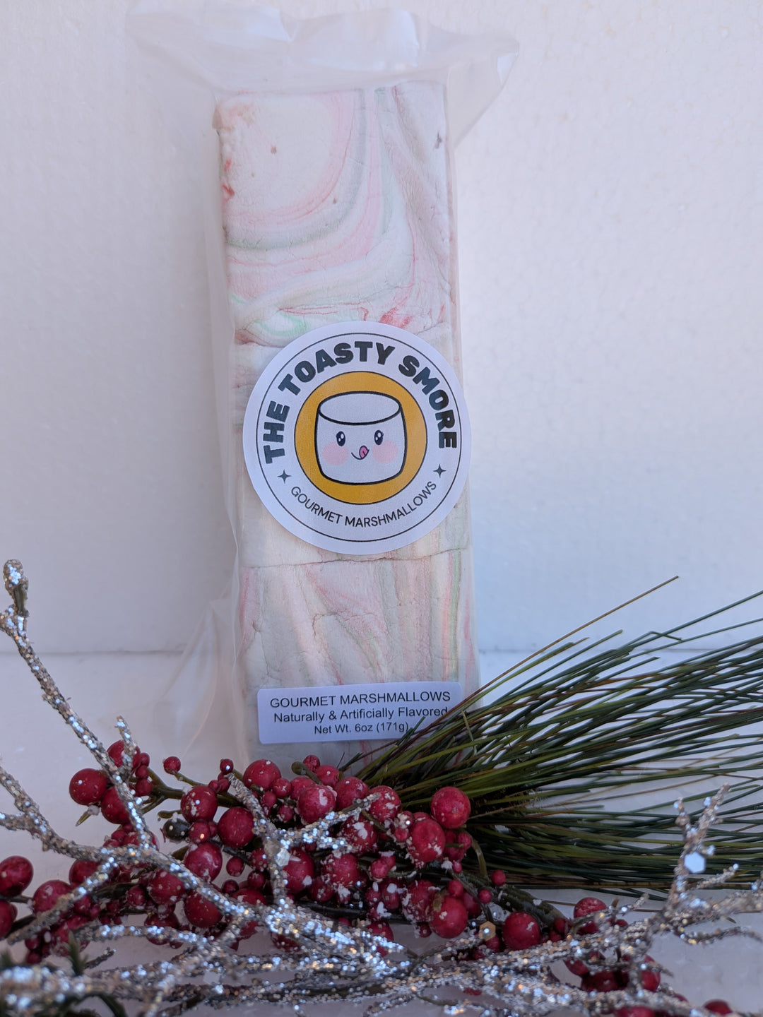 Packaged White Chocolate Peppermint Bark marshmallows by The Toasty S’more, styled with festive Christmas branches. Handcrafted, corn-syrup free, and made with beef gelatin and organic marshmallow root for a clean-label holiday indulgence.
