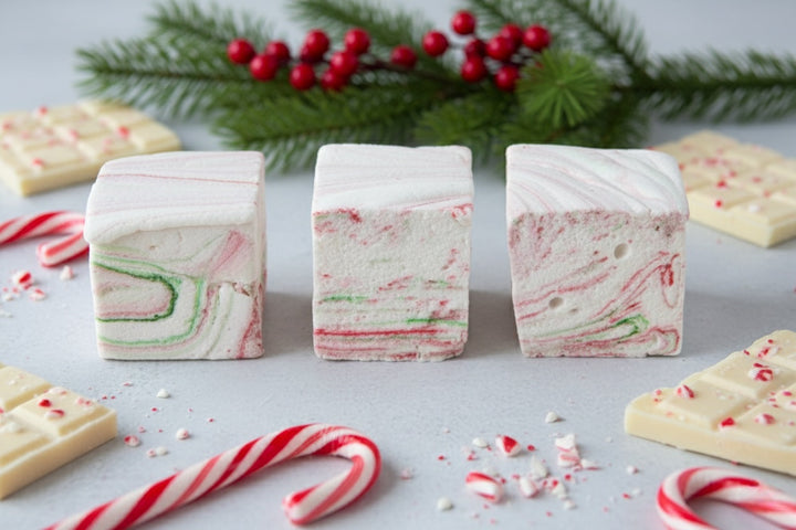 White Chocolate Peppermint Bark marshmallows by The Toasty S’more arranged in a neat row with crushed candy canes, white chocolate pieces, and festive Christmas branches on a bright white background. Handcrafted with beef gelatin and organic marshmallow root for a corn-syrup free, clean-label holiday indulgence.