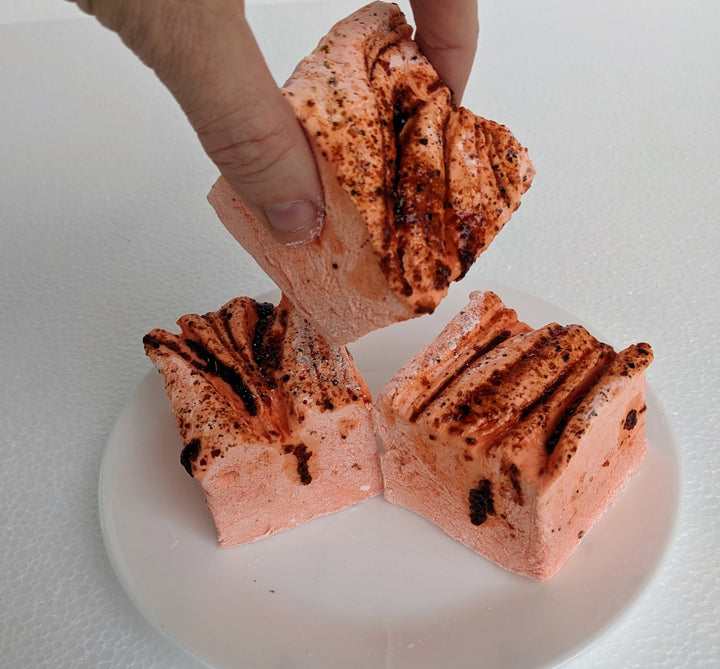 Three vibrant orange mango chamoy marshmallows on a white plate, displayed on a plain white background. A hand is seen picking up one of the marshmallows.