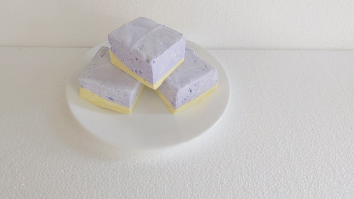 Watch a plate of three handcrafted gourmet lavender & honey marshmallows on a clean white background. A hand picks up one for a satisfying squeeze test, showcasing its soft, fluffy texture and delicate floral flavor.
