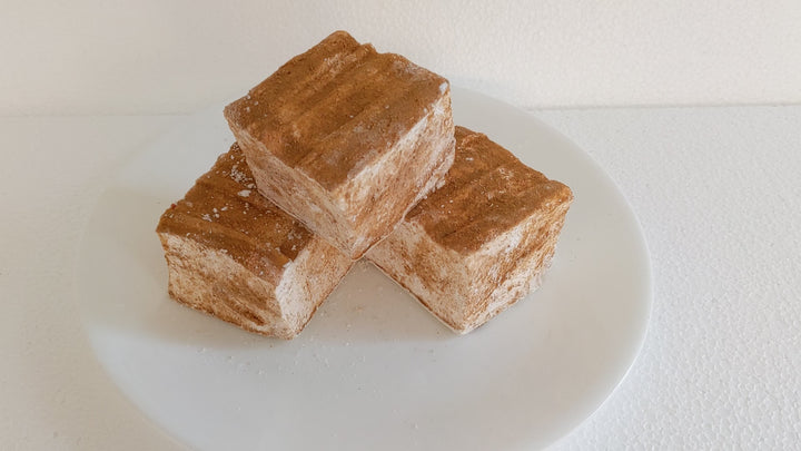 The video opens with a close-up shot of three caramel churro marshmallows on a white plate, displayed on a plain white background. A hand enters the frame, gently picking up one of the marshmallows and squeezing it to showcase its soft, fluffy texture. As the hand releases the marshmallow, it quickly returns to its original shape, highlighting its resilience and quality.