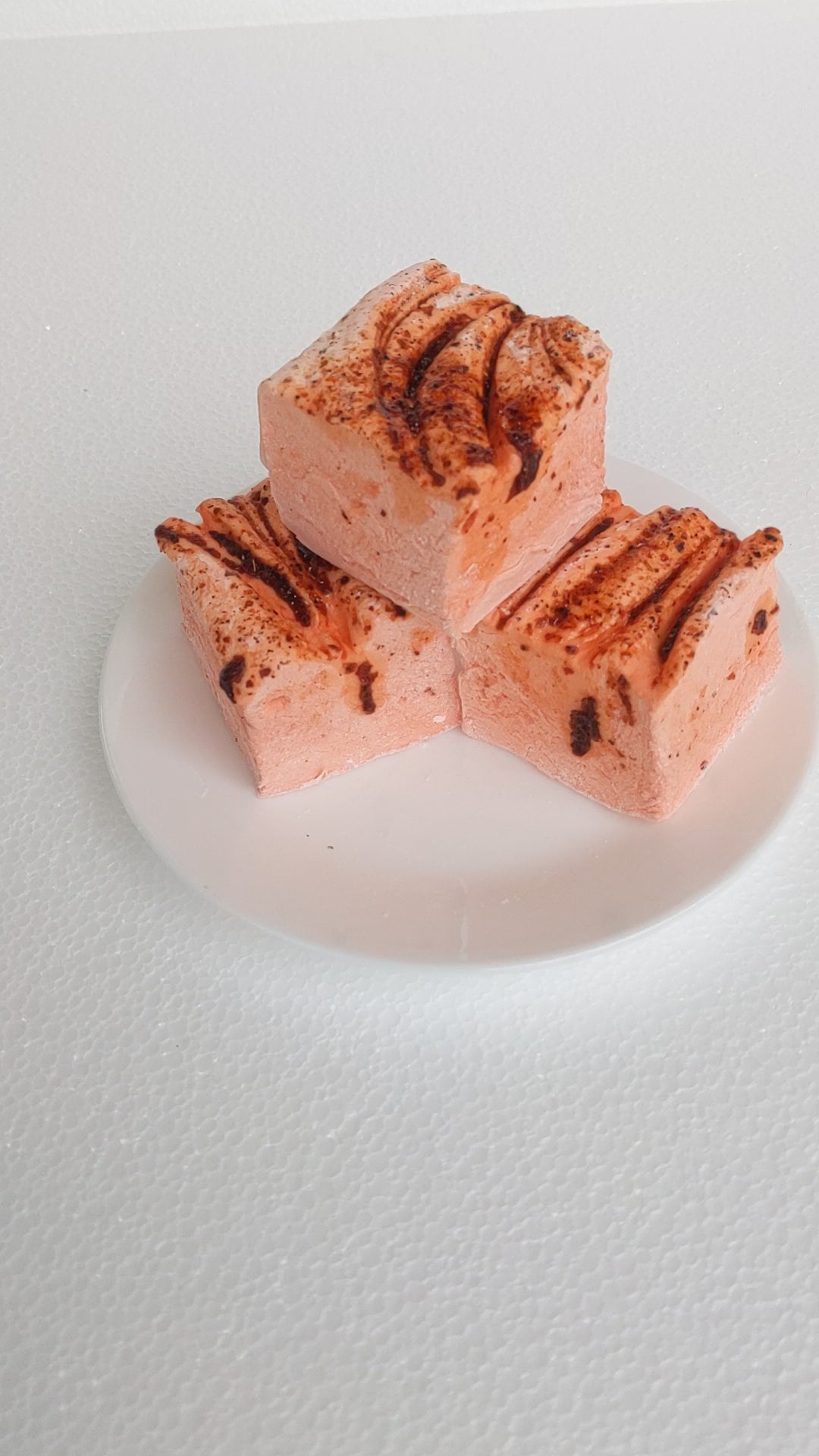 The video opens with a close-up shot of three vibrant orange mango chamoy marshmallows on a white plate, displayed on a plain white background. A hand enters the frame, gently picking up one of the marshmallows. The hand then squeezes the marshmallow, highlighting its soft and fluffy texture as it quickly returns to its original shape. The video emphasizes the marshmallows' handcrafted quality