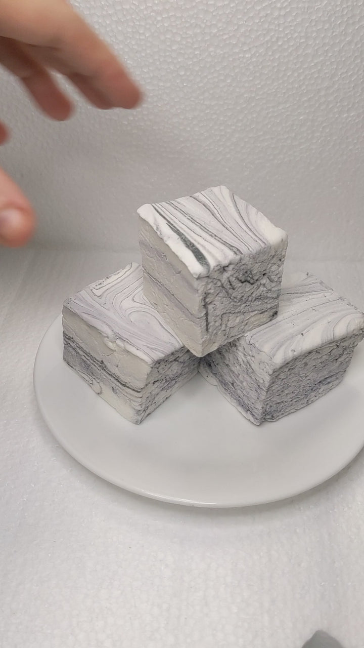 The video opens with a close-up shot of a single black licorice marshmallow, featuring a white base with elegant black swirls and streaks. The marshmallow is placed on a clean, white background, providing a crisp and clear view of its artisanal details. A hand slowly enters the frame and gently picks up the marshmallow, showcasing its soft and fluffy texture. As the hand squeezes the marshmallow, the camera captures the satisfying compression and bounce-back, highlighting its pillowy softness and gourmet qu