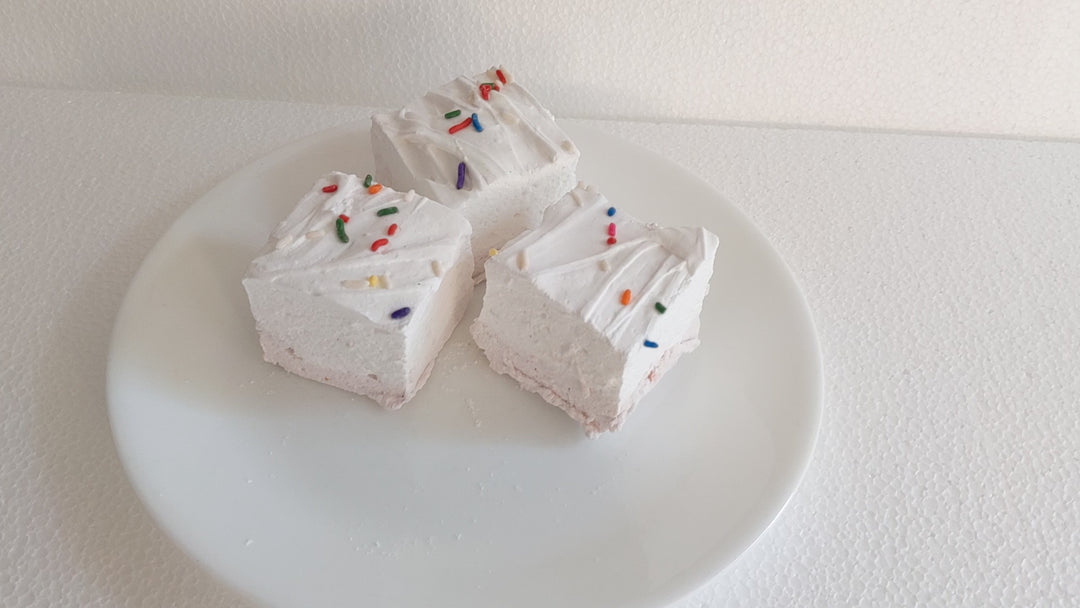 The video opens with a close-up shot of a single birthday cake flavored marshmallow, featuring a yellow bottom layer and a white top layer adorned with colorful sprinkles. The marshmallow is placed on a clean, white background. A hand slowly enters the frame and gently picks up the marshmallow, showcasing its soft, fluffy texture. As the hand squeezes the marshmallow, the camera captures the slight compression and bounce-back of the marshmallow, emphasizing its pillowy softness and artisanal quality. The vi