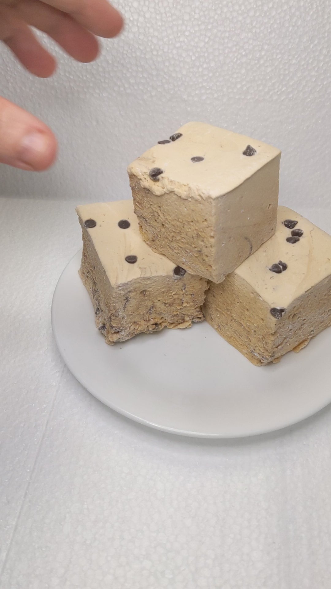 The video opens with a close-up shot of three cookie dough marshmallows on a white plate, displayed on a clean, white background. The marshmallows have a soft, fluffy texture with a delightful cookie dough flavor. A hand slowly enters the frame and gently picks up one of the marshmallows, showcasing its pillowy softness and detailed artisanal quality. As the hand squeezes the marshmallow, the camera captures the satisfying compression and bounce-back, emphasizing the gourmet nature of this unique treat. The