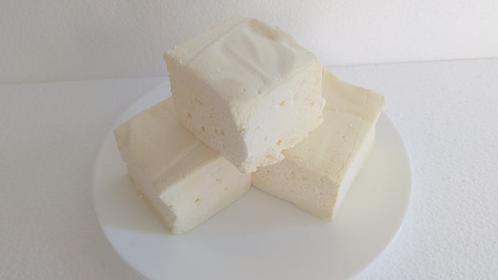 The video begins with a close-up shot of a single yellow butter vanilla marshmallow on a white plate, displayed on a plain white background. A hand enters the frame, gently picking up the marshmallow. The hand then squeezes the marshmallow, highlighting its soft and fluffy texture as it quickly returns to its original shape.