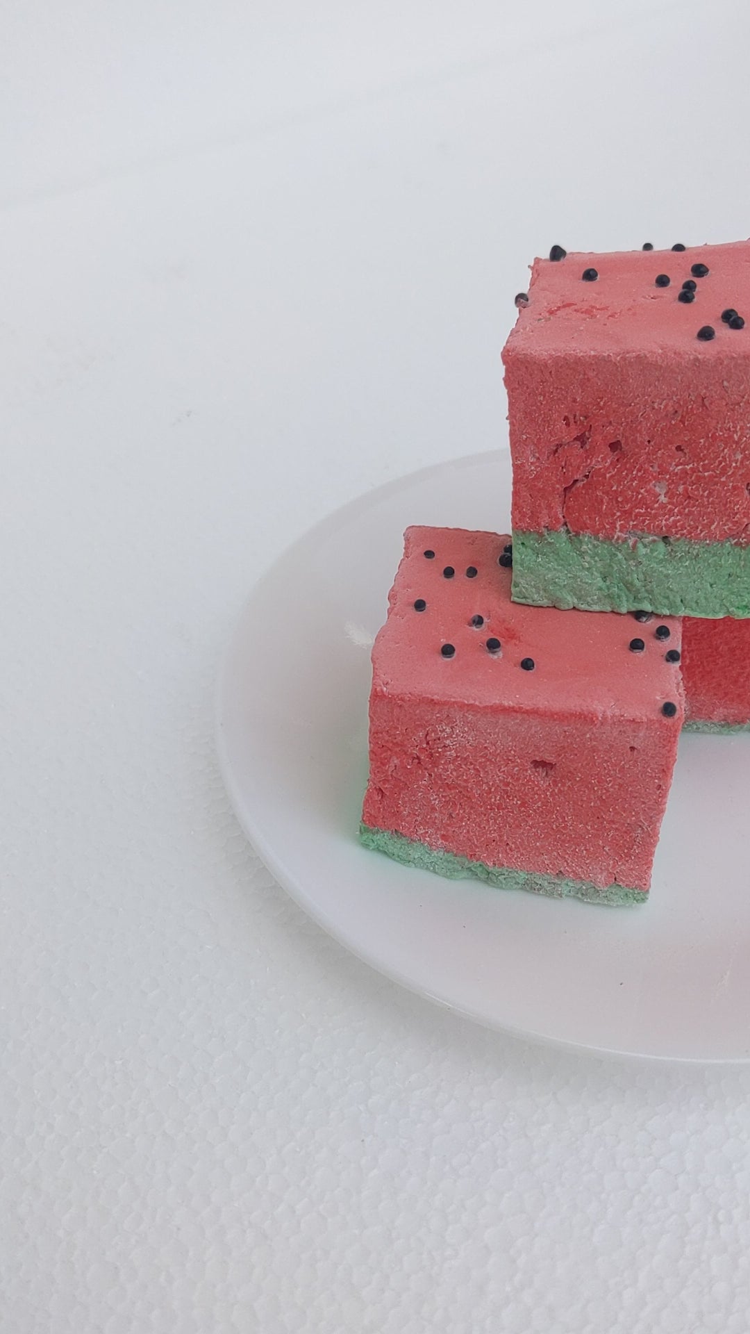 A close-up view of three of the best fruity marshmallows, inspired by watermelon, arranged on a white plate. Each marshmallow features a green base and a red top, designed to resemble a juicy watermelon slice. Tiny black sprinkles are scattered across the red layer, mimicking watermelon seeds. The bright colors contrast beautifully against the clean, white plate, emphasizing their fluffy texture and handcrafted quality. A fun and delicious treat for fruit-flavored marshmallow lovers!