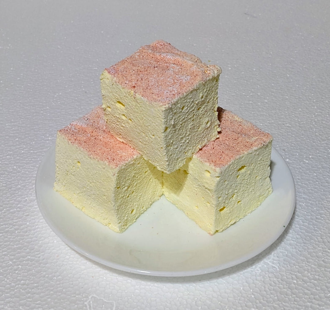 Three soft, 2-inch pink lemonade gourmet marshmallows with pale pink and soft yellow swirls rest on a white plate, their pastel hues popping against the bright background.