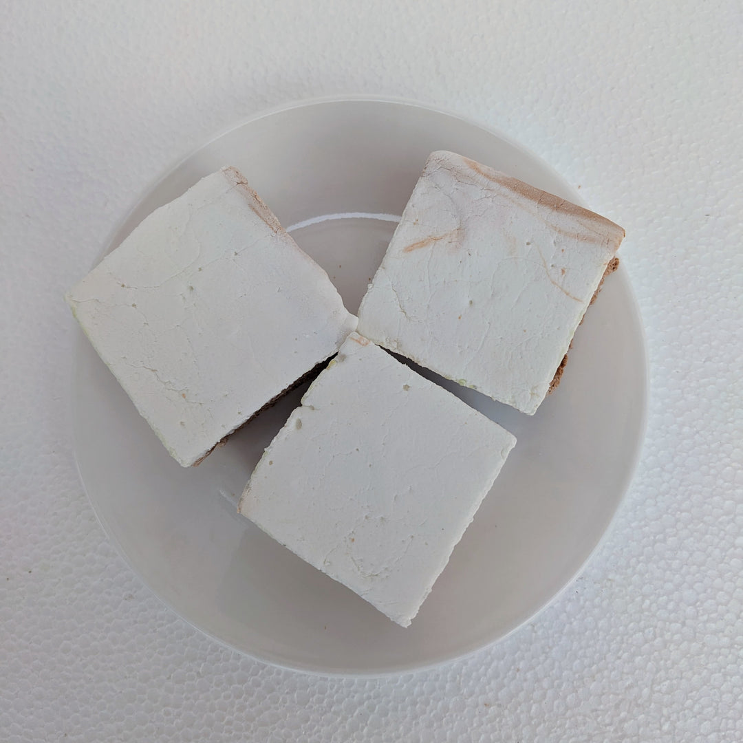 Top-down view of three jumbo Buttered Beer gourmet marshmallows with golden butterscotch shimmer on a white plate, one slightly forward to highlight its pillowy texture. Handcrafted in Texas with organic marshmallow root, beef gelatin, and no corn syrup, these 2-inch cubes are gluten free and perfect for s’mores, desserts, or clean-label gifting.